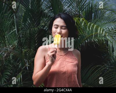 Woman eating popsicles. Happy beautiful Asian woman wearing casual pink tank top holding yellow popsicle on green tropical palm leaves background, out Stock Photo