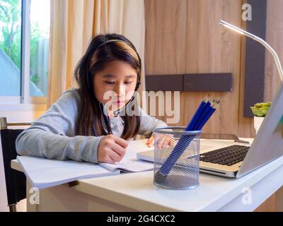 Asian girl in headphones sit at desk study online on laptop. Kid wear headset handwriting in notebook learning using internet lessons on quarantine. G Stock Photo