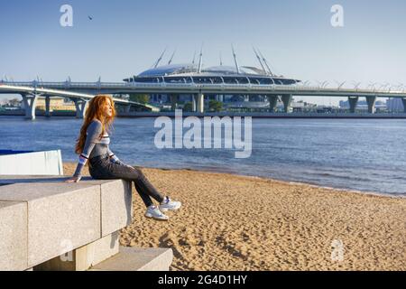 Beautiful young woman with long hair of Caucasian nationality , in casual clothes sits on the embankment of the river on a spring sunny day with a view of the bridge Stock Photo