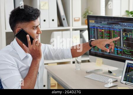 Trader pointing at computer screen with stock market data when talking on phone with client or colleague Stock Photo