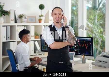 Cheerful experienced trader talking on phone with client and smiling at camera, his colleague working in background Stock Photo