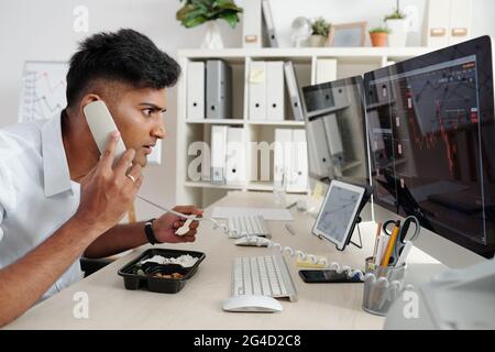 Excited trader looking at ascending chart on screen with stock market data and making phone call to colleague when eating lunch at his desk Stock Photo