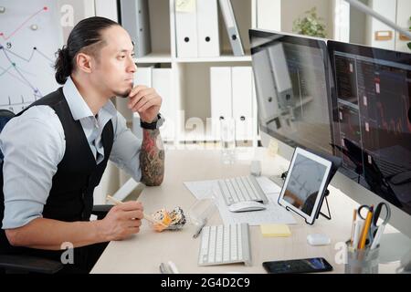 Pensive trader eating rolls when analyzing stock market trends on computer screen in front of him, thinking what stocks and futures to buy and sell Stock Photo