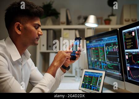 Serious broker checking stock soar via application on smartphone when working in dark office Stock Photo