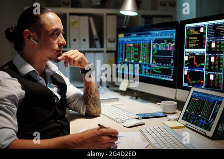 Pensive serious trader with man bun looking at scren with charts and shares prices and taking notes on paper Stock Photo