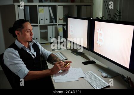 Pensive serious investor looking at inscreasing bitcoin prices chart and writing on sticking notes Stock Photo