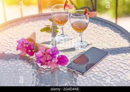 Glasses with homemade liqueur, decorative with fresh summer surreal flowers and mobile phone. Feminine flat lay. Summer bright background for advertis Stock Photo