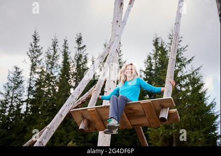 Low angle of beautiful young woman looking at camera and smiling while swinging on wooden swing in coniferous forest. Concept of playfulness, fun and active leisure. Stock Photo
