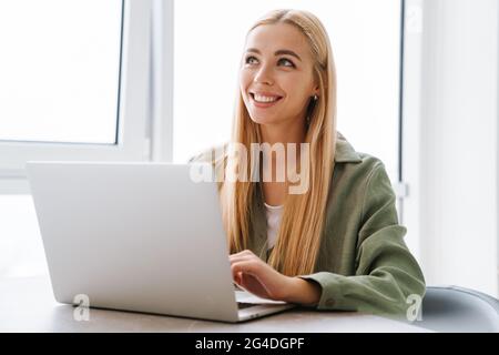 Happy attractive young blonde woman sitting at the table with laptop computer indoors Stock Photo