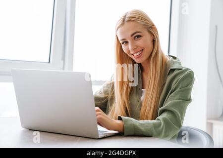 Happy attractive young blonde woman sitting at the table with laptop computer indoors Stock Photo