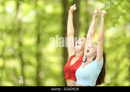 Two excited friends celebrating vacation raising arms in a green forest or park
