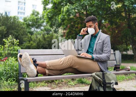 Young man with respirator using laptop and smartphone outdoors in city, covid-19 concept. Stock Photo