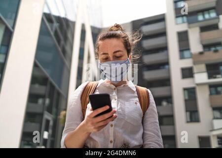 Portrait of young woman with face mask using smartphone outdoors in city, life after covid-19 vaccination. Stock Photo