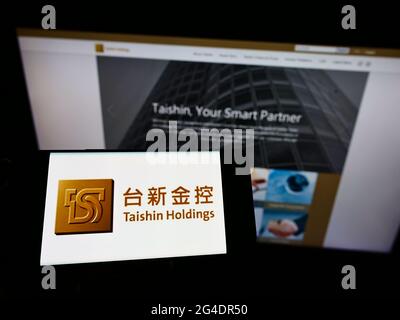 Person holding cellphone with logo of Tainwanese company Taishin Financial Holding Co. Ltd. on screen in front of webpage. Focus on phone display. Stock Photo