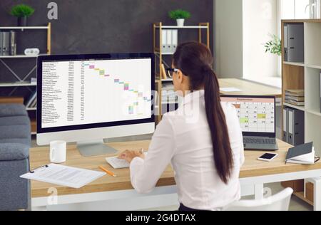 Back view of woman sitting at office desk and working on desktop computer and laptop Stock Photo