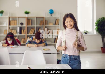 Happy smiling girl pupil standing with backpack next to classroom desk portrait Stock Photo