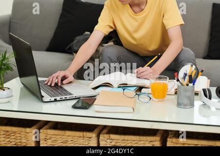 Cropped image of schoolboy doing homework at home, he is checking article on laptop screen and underlining sentences in students book Stock Photo