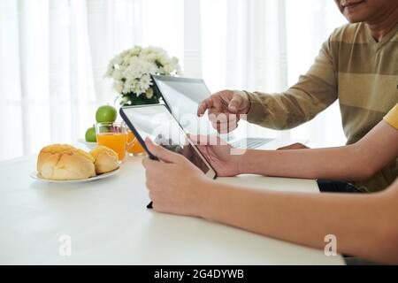 Father helping teenage son to install application for online studying on tablet computer when they are sitting at kichen table Stock Photo