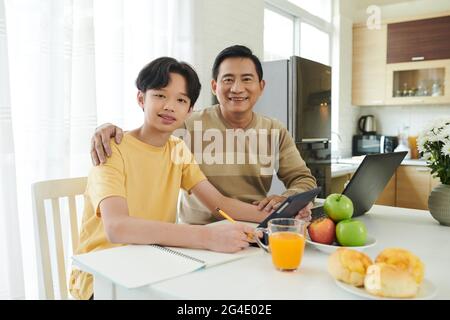 Portrait of happy hugging father and son sitting at kitchen table when working and doing homework Stock Photo