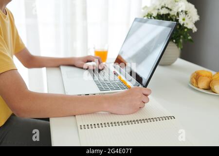 Hands of schoolboy reading book or article on laptop and writing in textbook at desk in his room Stock Photo