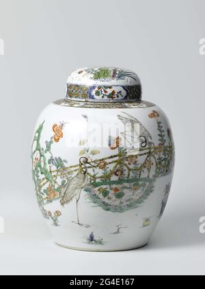 . Egg-shaped porcelain lid pot, painted on the glaze in blue, red, green, yellow, eggplant and black. On the wall a fenced garden with two cranes, one standing and one on the fence, at a pond with lotus plants, a rock and a flowering tree (Magnolia). To the pond flowering plants (Chrysanthemum, Prachtanjer, Peony, Aster, Lily) and two flower pots with pine, bamboo and orange. In the Magnolia tree a parrot on a rack. In addition to two flying birds. On the shoulder napkin interspersed with flower branches in cartouches. The lid is later in the same style with underglaze blue advanced from Delft Stock Photo