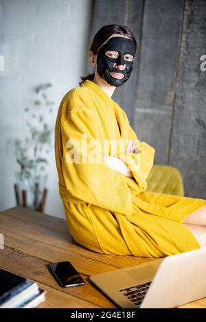 Woman in yellow bathrobe with black cleansing mask on her face at home office Stock Photo