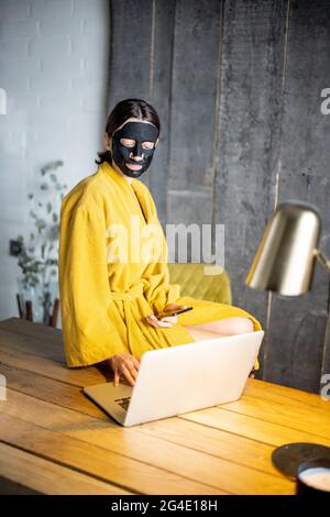 Woman in yellow bathrobe with black cleansing mask on her face at home office Stock Photo