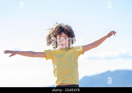 smiling boy with long hair jumps and has fun playing with open arms. blue sky background on a sunny day Stock Photo