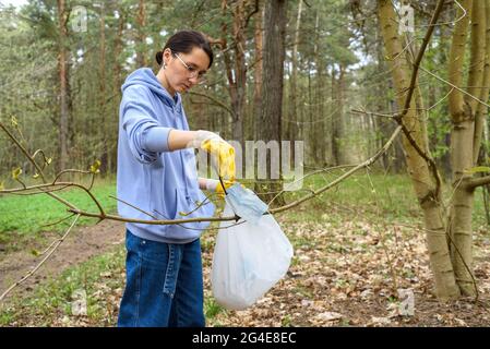 Woman in casual wear, gloves, with plastic trash bag collecting garbage in forest. Plogging Stock Photo