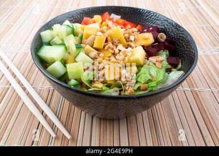 Vegetarian poke bowl in design black bowl with chopsticks below, on a reed pad background. Green cucumber, red pepper, peanuts, corn, beetroot on top Stock Photo