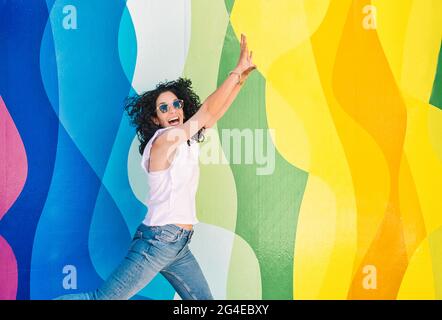 Young brunette woman with curly hair, jeans and sunglasses has fun while jumping on a colorful wall. leisure and free time concept Stock Photo