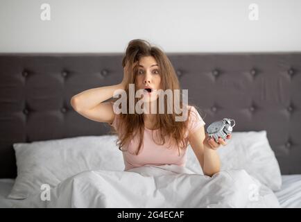 Terrified yound woman holding alarm clock in bed at home, grabbing her head, oversleeping to work Stock Photo