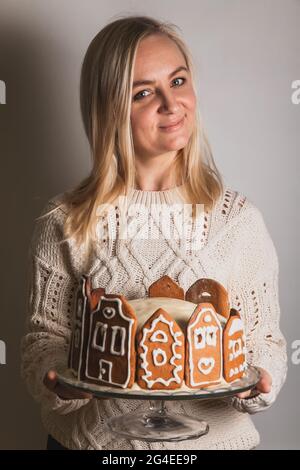 Woman holds homemade cake with gingerbread city house facades, a Christmas tree and reindeer on glass stand Stock Photo