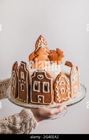 Woman holds homemade cake with gingerbread city house, Christmas tree and reindeer on glass stand, with copy space. Stock Photo