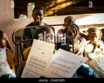 MBOUR, SENEGAL - DECEMBER, Circa, 2020. Unidentified group of cheerful children sitting on chairs outdoors, looking at teacher to learn song with musi Stock Photo