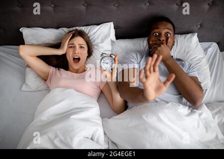 Top view of stressed interracial couple with alarm clock shouting in terror, being late for work, lying in bed, top view Stock Photo