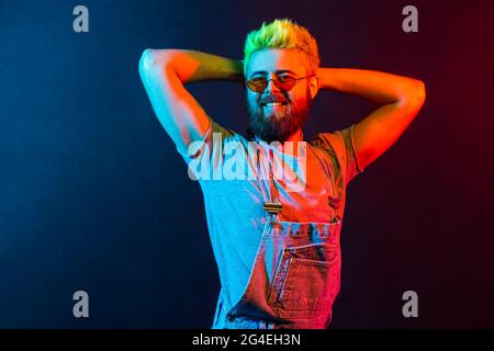 Happy bearded hipster man with green hair posing with satisfied facial expression, has confident look, raising hands behind his head, dresses overalls Stock Photo