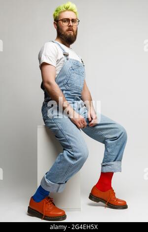Full length photo of confident concentrated man wearing jacket, denim overalls and stylish red shoes, looking at camera with serious expression. Indoo Stock Photo