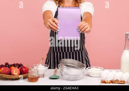 Cropped image of woman wearing black striped apron holding and showing blank paper notebook to camera, bakery products on table, cooking cakes. Indoor Stock Photo