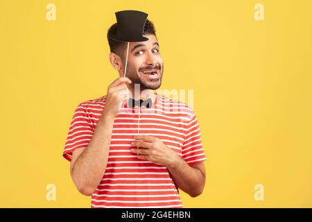 Happy satisfied man with beard in striped t-shirt enjoying having fun trying on paper mask on stick with hat and bow tie, preparing for masquerade. In Stock Photo