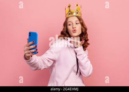 Portrait of confident curly haired teenage girl in hoodie with crown sending air kiss on cellphone camera, modern technology, selfie. Indoor studio sh Stock Photo