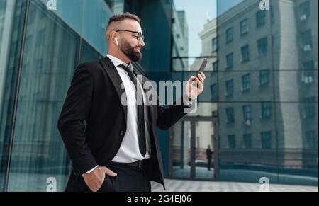 Happy businessman in suit talking on smartphone in wireless earphones standing near glass office building outdoors Stock Photo
