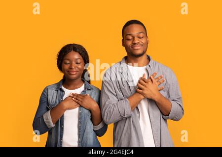 Portrait Of Calm Black Couple Keeping Hands On Chest, Feeling Blessed Stock Photo