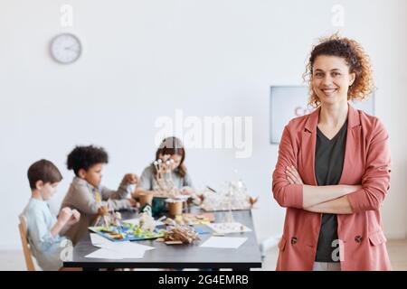 Waist up portrait of smiling female teacher looking at camera with group of children making wooden models during art and craft class in school, copy s Stock Photo