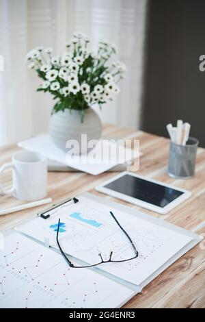 Vase with flowers, folder with reports, glasses and tablet computer on table of person working from home Stock Photo