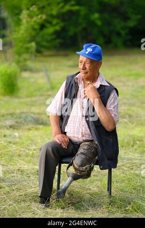 Disabled old man with a vintage wooden prosthetic limb sitting on a bench on a lawn Stock Photo