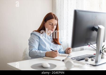 Happy stylish woman remote working from home distance office on pc taking notes. Smiling business lady using computer watching webinar Stock Photo