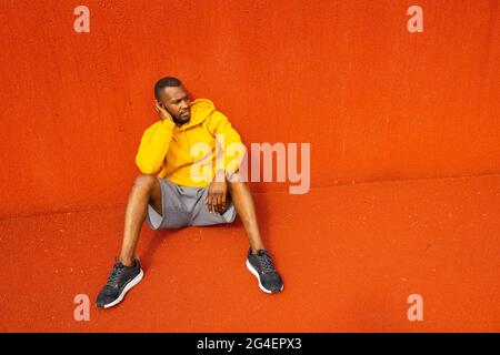 Tired pensive athlete sportsman sitting isolated near red wall, having rest after workout, training, game, match. Serious African-American male in Stock Photo