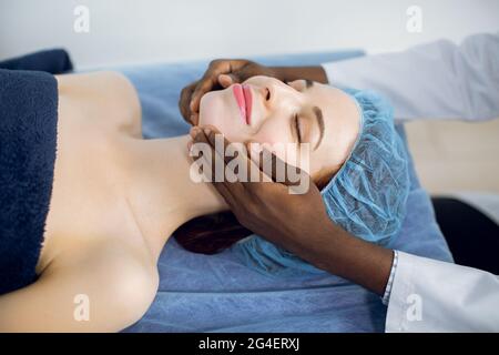 Relaxing treatment in medicine. Caucasian woman in disposable cap, lying on the couch, enjoying facial massage with closed eyes in spa salon or clinic. Hands of male black chiropractor doing massage Stock Photo