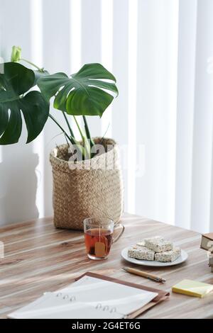 Monstera plant, planner, plate with delicous corn bars and cup of tea on table in apartment Stock Photo